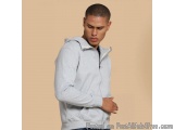 The image shows a young man wearing a light grey hoodie. He is standing in front of a plain beige background and is looking d...