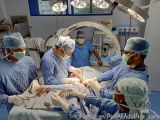 The image shows a group of surgeons performing an operation in an operating room. There are six surgeons in the image, all we...