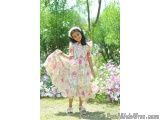 The image shows a young girl standing in a garden with a beautiful floral dress. She is wearing a white headband with pink fl...