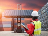 The image shows a man wearing a white hard hat and a yellow high visibility vest, standing in front of a large warehouse with...