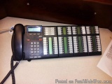 The image shows a black telephone on a desk. The telephone has a corded design and a large display screen. The display screen...
