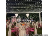 The image shows an outdoor event space with a pergola covered in string lights. The pergol is decorated with white and pink s...