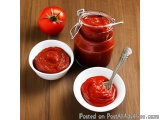 The image shows a jar of tomato sauce with a silver spoon in it. The jar is made of glass and has a metal lid. Next to the ja...