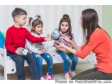 The image shows a mother and her four children sitting on a white couch in a living room. The mother is sitting on the floor...