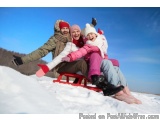 The image shows a family of three - a man, a woman, and a young girl - sledding down a hill in the snow. They are all wearing...