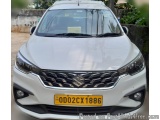 The image is a photograph of a white Maruti Suzuki car. The car is parked on a street with a concrete wall and some plants in...