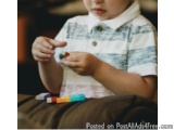 The image shows a young boy sitting on a couch and playing with colorful crayons. He is wearing a white collared shirt with a...