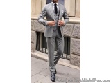 The image shows a man walking on a sidewalk. He is wearing a grey suit with a white shirt and a black tie. The suit has a sin...