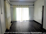 The image shows an empty room with wooden flooring and white walls. The room has a large window with sheer white curtains han...