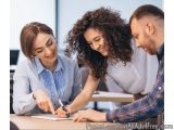 The image shows three people, two women and a man, sitting at a desk in an office. They are all smiling and appear to be enga...
