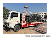 The image shows a white truck with a red stripe on the side. The truck is parked on a dirt road with a building in the backgr...