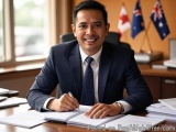 The image shows a man in a suit sitting at a desk in an office. He is smiling and holding a pen in his hand and appears to be...