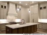This image is of a modern kitchen with a large island in the center. The island has a white countertop and a sink with a blac...