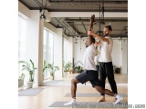 The image shows two men in a yoga studio. They are both wearing white t-shirts and black shorts and are standing on gray yoga...