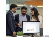 The image shows three people in an office setting, two men and a woman, standing in front of a computer monitor. The man on t...