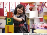 The image shows a young girl, probably around 4-5 years old, standing in a classroom. She is wearing a black and red plaid dr...