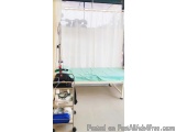 The image shows a hospital room with a single bed in the center. The bed is covered with a green sheet and there is a medical...