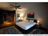The image shows a modern and spacious bedroom with a large bed in the center. The bed has a black headboard and white linens,...