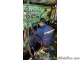 The image shows a man working on a large industrial machine in a workshop. He is wearing a blue uniform with the words "PA Po...