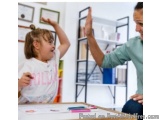 The image shows a young girl and a woman sitting at a table in a classroom. The girl is wearing a white t-shirt with the word...
