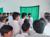 The image shows a group of young men gathered in a room with a green curtain in the background. They are all wearing white sh...