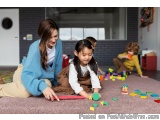 The image shows a mother and her two young children playing with colorful building blocks on the floor in a classroom. The mo...