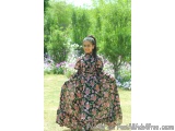 The image shows a young girl standing in a garden. She is wearing a long, flowing dress with a floral pattern in shades of pi...
