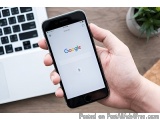 The image shows a person's hand holding a black iPhone with the Google logo on the screen. The phone is resting on a wooden d...