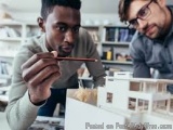 The image shows two young men in an office setting, looking at a model of a building. The man on the left is holding a pair o...