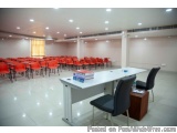 The image shows a large conference room with rows of red chairs arranged in neat rows. The room has a white tiled floor and a...
