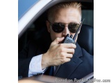 The image shows a man in a suit and sunglasses sitting in a car. He is holding a walkie-talkie in his right hand and appears...