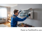In this image, we can see a man working on an air conditioning unit in a living room. He is wearing a blue uniform and gloves...