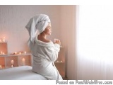 The image shows a woman sitting on a bed with a white towel wrapped around her head. She is holding a cup of coffee in her ha...
