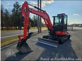 The image shows a red mini excavator parked on the side of a road. The excavator has a long arm with a bucket attached to it,...