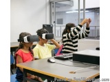 The image shows three children sitting at a desk in a classroom. They are all wearing virtual reality (VR) headsets and appea...