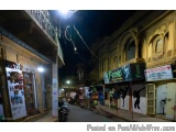 The image shows a street at night in an Indian city. The street is lined with shops and buildings on both sides, and there ar...
