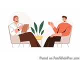 The image shows two people sitting in a room with a plant in the corner. The person on the left is a man wearing a white shir...