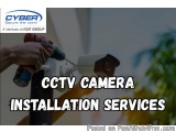 The image shows a person's hand holding a power drill and installing a CCTV camera on a pole. The background is blurred, but...