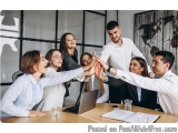 The image shows a group of young business people in a meeting room. They are gathered around a wooden table with a laptop and...