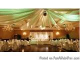 The image shows a large ballroom with a high ceiling decorated with green and white drapes and chandeliers. The room is set u...