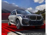 The image shows a silver BMW X1 SUV driving on a red and white striped road. The car is in motion, with the wheels slightly b...