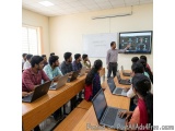 The image shows a classroom with a group of students sitting at desks with laptops in front of them. The desks are arranged i...