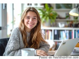 The image shows a young woman sitting at a desk with a laptop in front of her. She is smiling and looking at the camera. She...