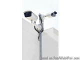The image shows a security camera mounted on a metal pole. The pole is attached to a white wall with a white building in the...