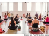 The image shows a group of people sitting on yoga mats in a large room with large windows. They are all facing the same direc...