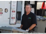 The image shows a man standing in front of a white truck with a warning sign on the side. He is wearing a black polo shirt wi...