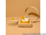 The image shows a small dessert on a wooden stand. The dessert is a round cake with a white base and a yellow top. It has a d...
