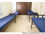 The image shows a row of four single beds in a room with white walls and a wooden door in the background. The beds are arrang...