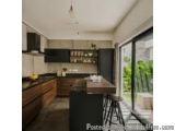 The image shows a modern kitchen with a large window on the right side. The kitchen has a large island in the center with a w...