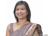 The image is a portrait of a middle-aged Indian woman. She is wearing a traditional Indian saree in a light purple color with...